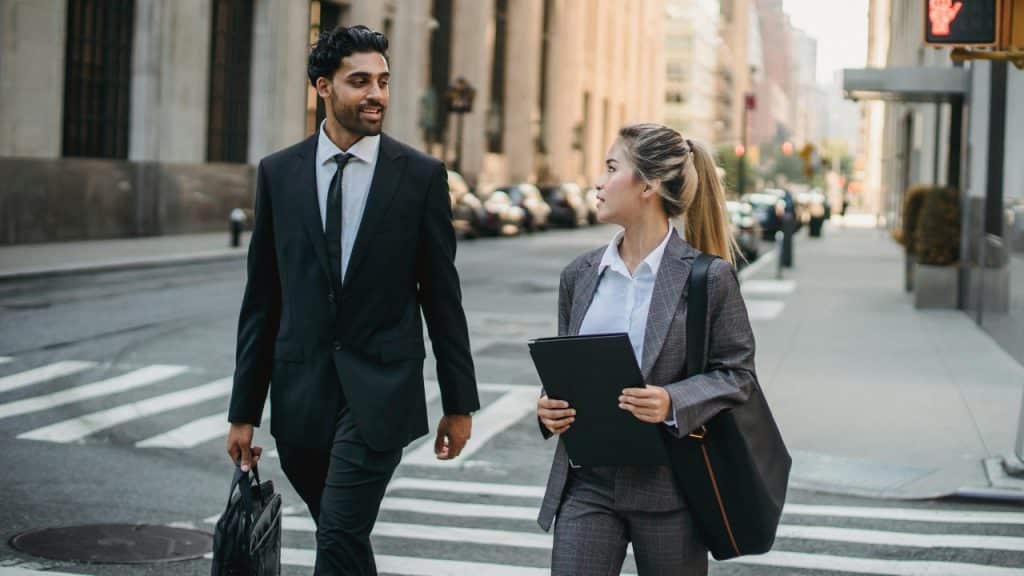 A man and woman in business attire walk across a city street while talking together.