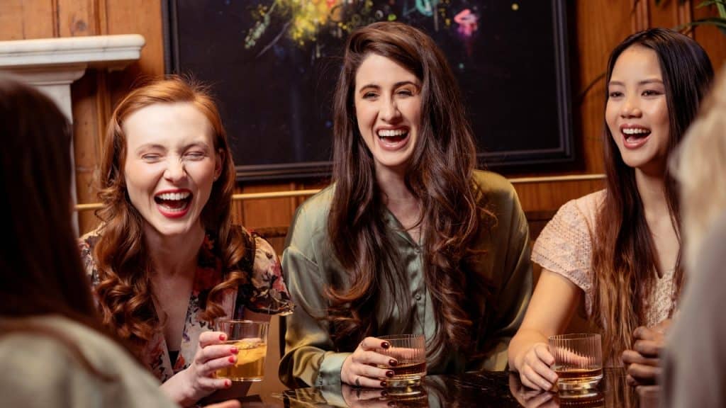 Three women sit at a dark table laughing together while holding glasses with amber liquid.