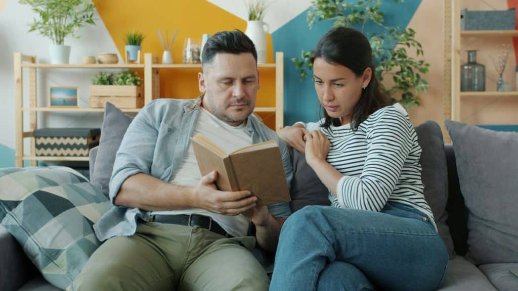 A man and a woman sit together on a sofa while reading a book.