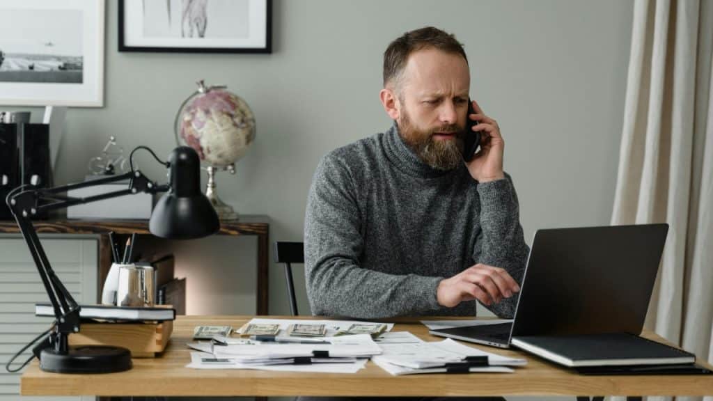 A bearded man on a phone call sitting at a desk with cash and a laptop.
