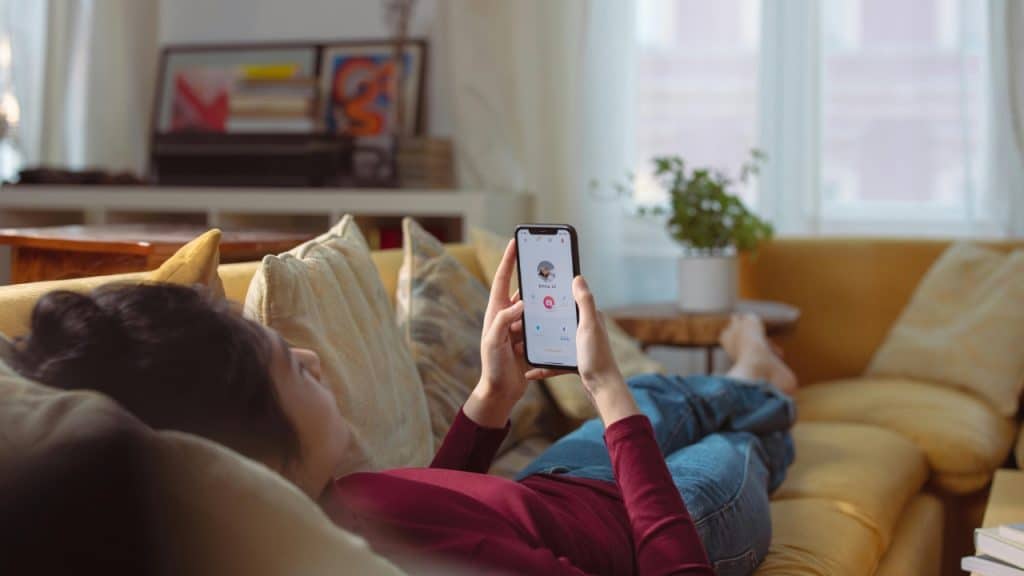 A person lies on a yellow sofa while holding a smartphone displaying a dating profile.