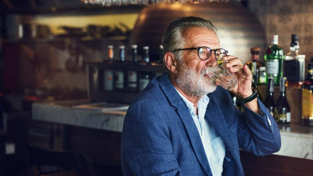 An older man with glasses and a blue blazer drinks from a glass at a bar.