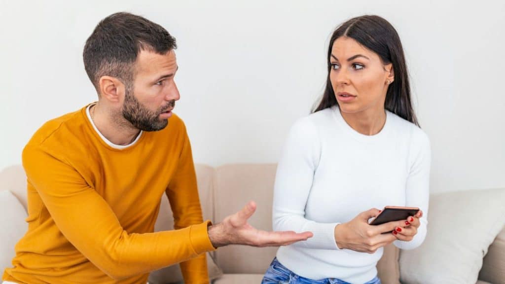 A man gestures toward a woman holding a phone, who looks back with a skeptical expression.