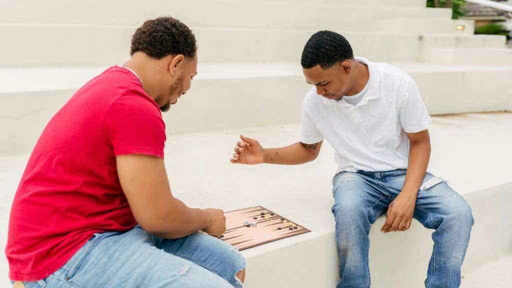 Two men sit on outdoor concrete steps playing a board game with dice and tokens