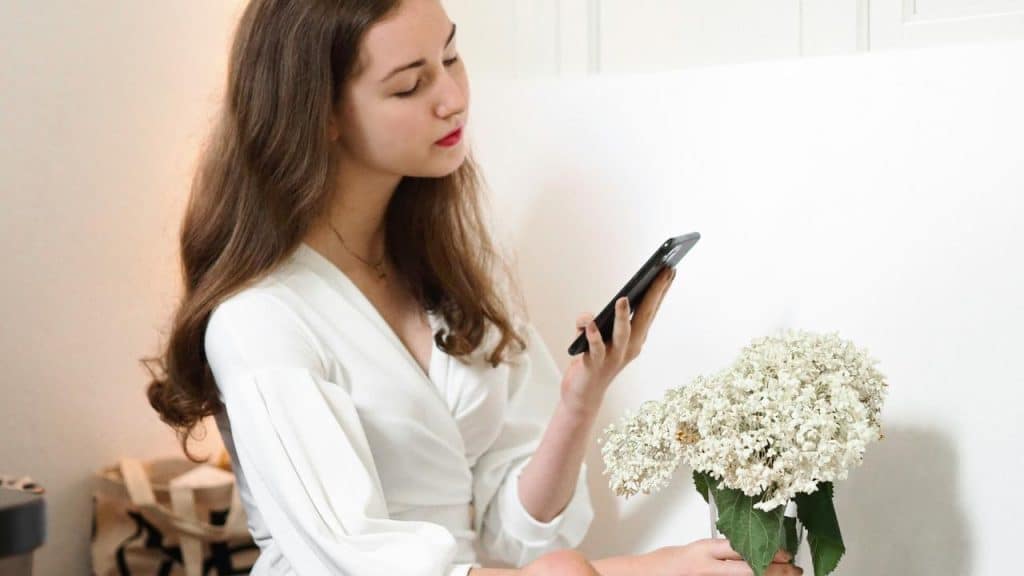 A woman holding a smartphone while sitting beside a bouquet of white flowers.