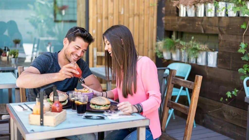 A man and woman smiling while adding ketchup to fries at an outdoor café.