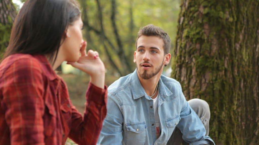 A man in a denim shirt speaks to a woman sitting near a large tree.