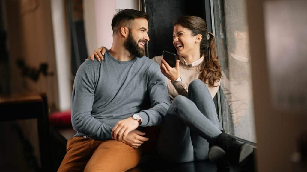 A man and woman sitting on a window ledge laughing while looking at a smartphone.