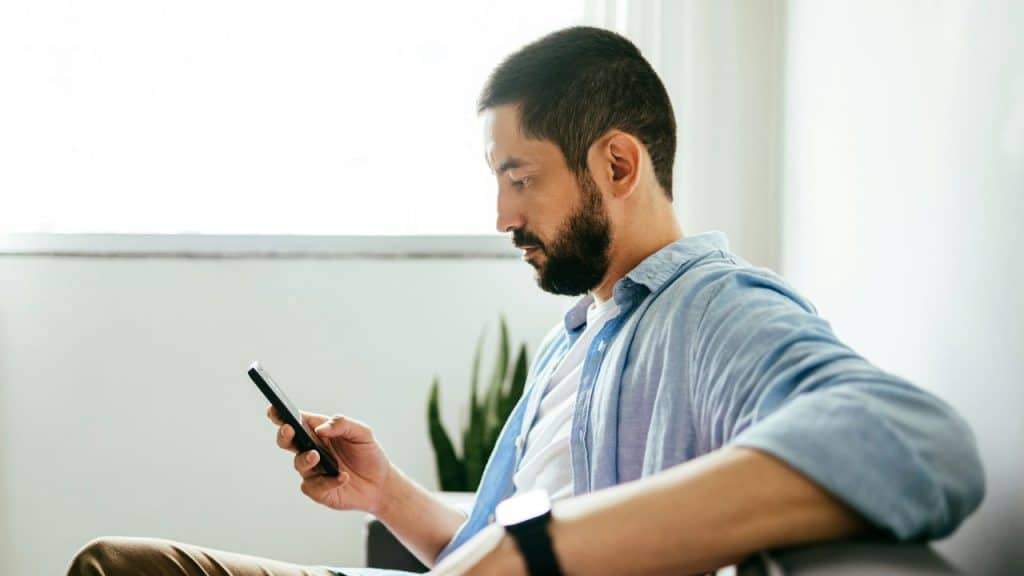 A bearded man in a blue shirt sits indoors looking intently at his handheld smartphone.
