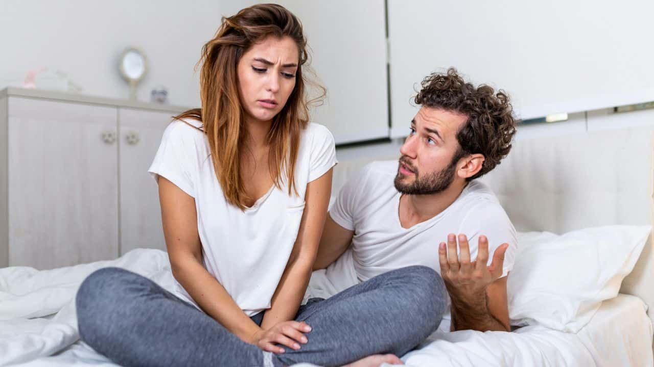 A woman sits cross-legged on a bed while a man gestures and speaks to her.