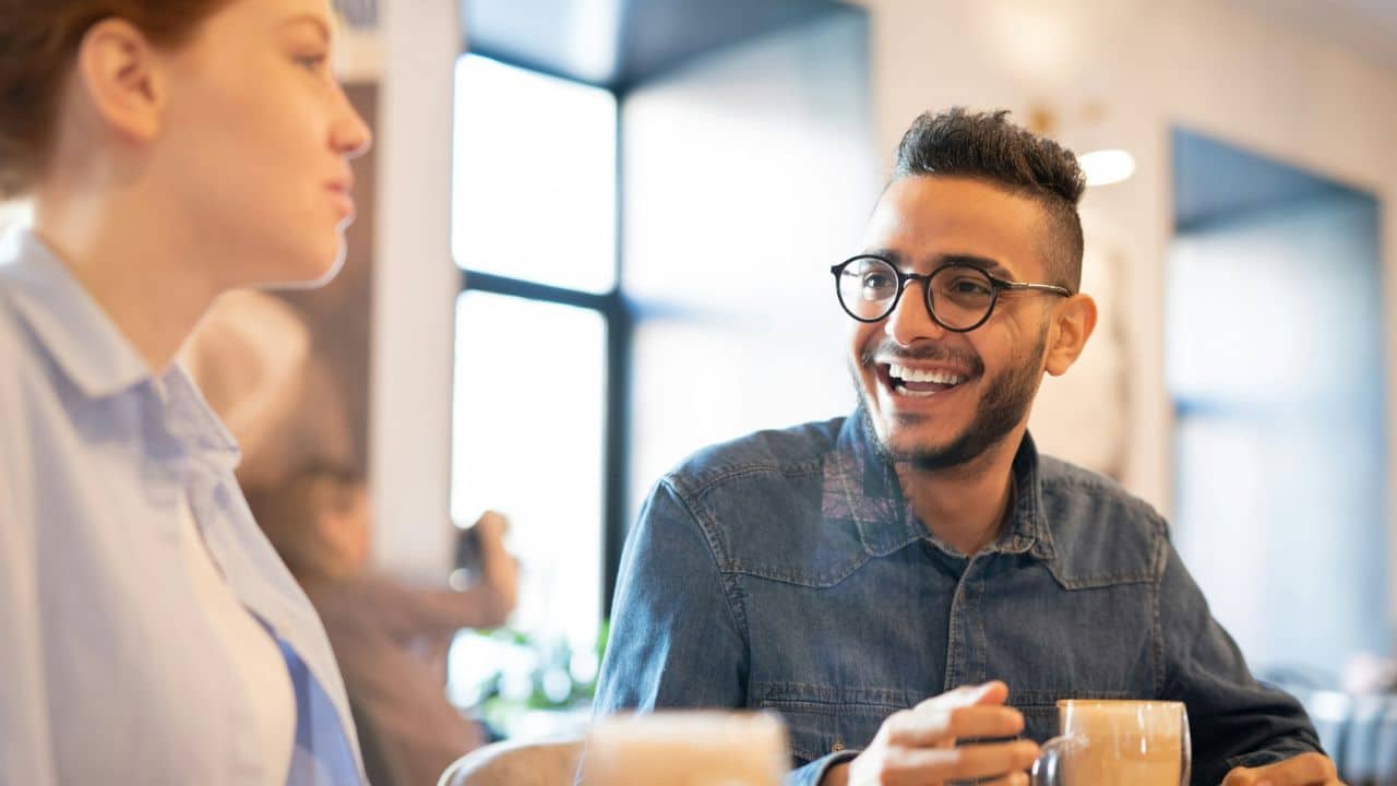 A smiling man wearing glasses speaks to a woman while sitting in a bright cafe.