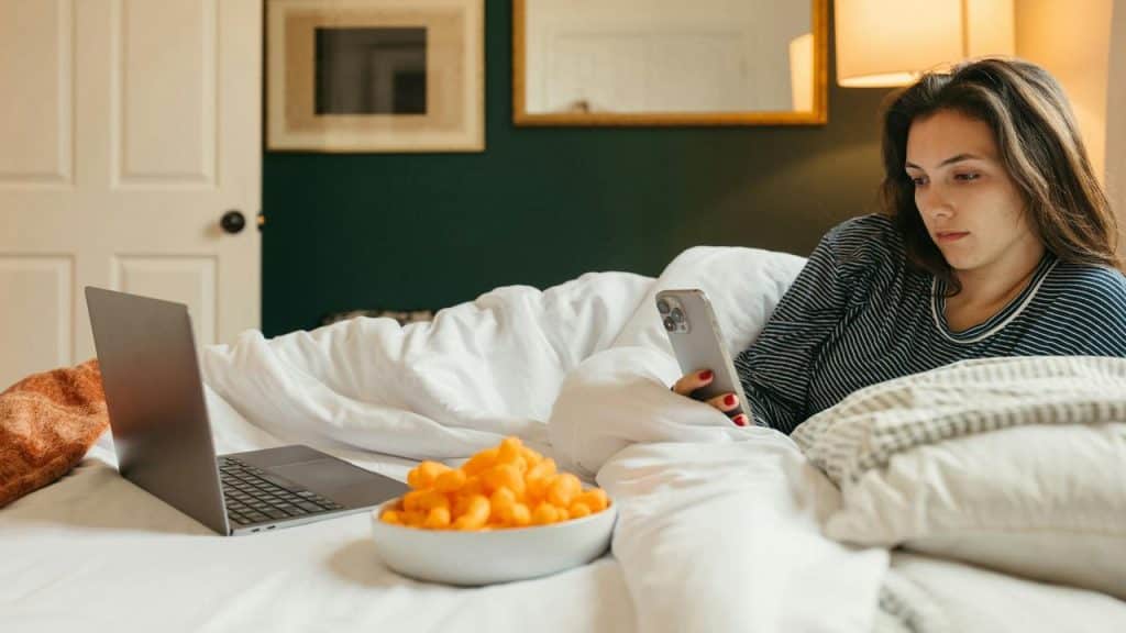 A woman lying in bed, looking at her phone beside a laptop and snack bowl.