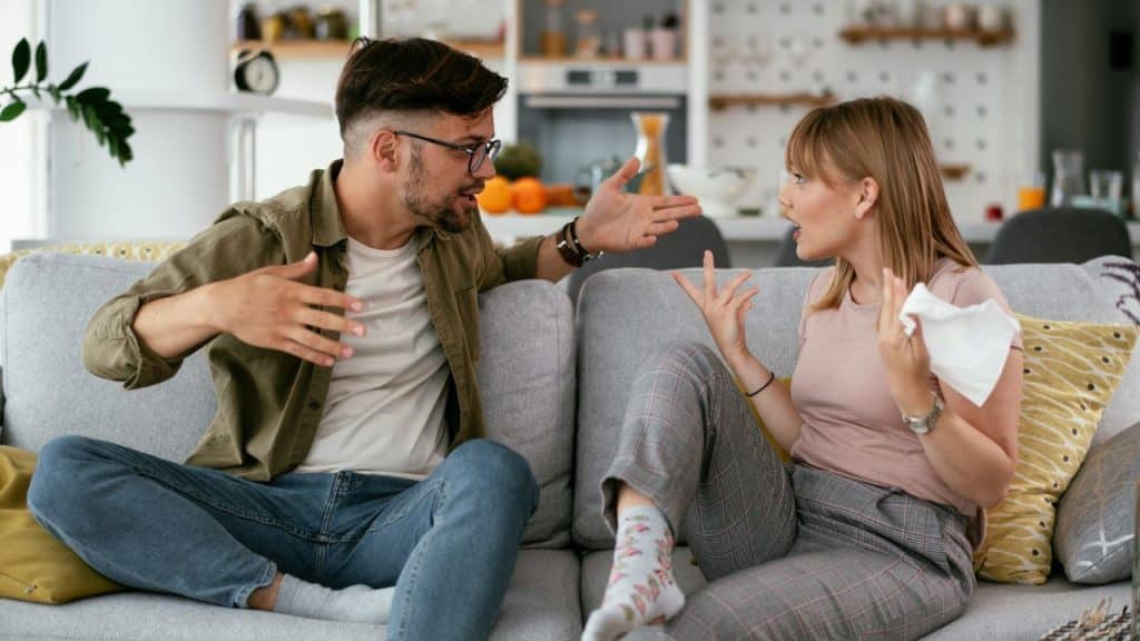 A man and woman sit on a gray sofa facing each other with raised hands.