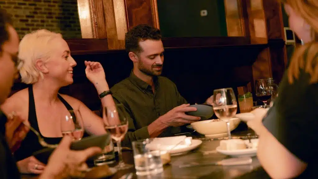 Four people sit around a long dinner table with food, wine glasses, and warm lighting.