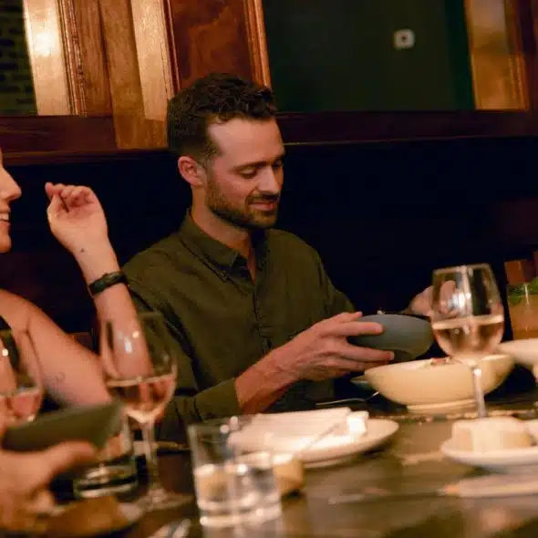 Four people sit around a long dinner table with food, wine glasses, and warm lighting.
