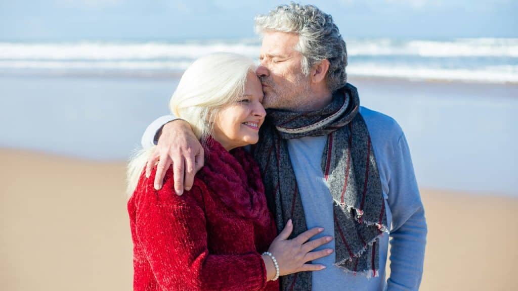 An older man kisses the forehead of an older woman while standing on a beach.