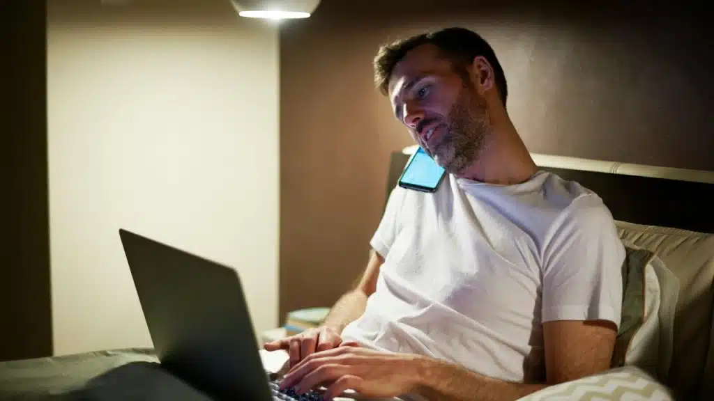 A man in a white t-shirt sits in bed using a laptop and a mobile phone.