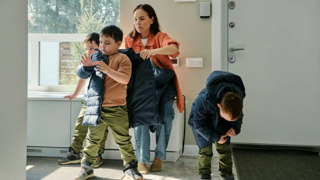 A woman helps a child into a jacket while two other children stand in a room.