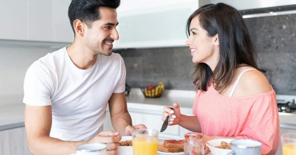 A couple smiling at each other while having breakfast in a kitchen.