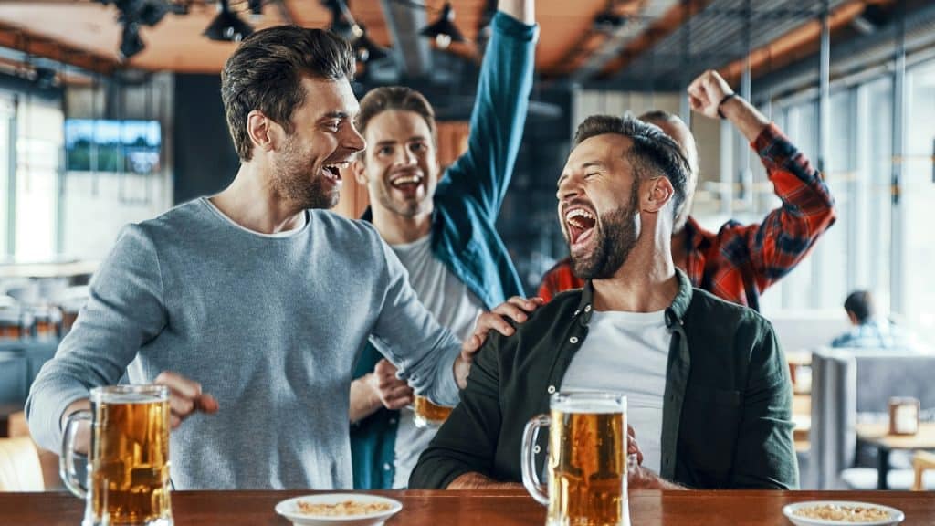 Four men cheer and laugh at a bar with mugs of beer on the counter.