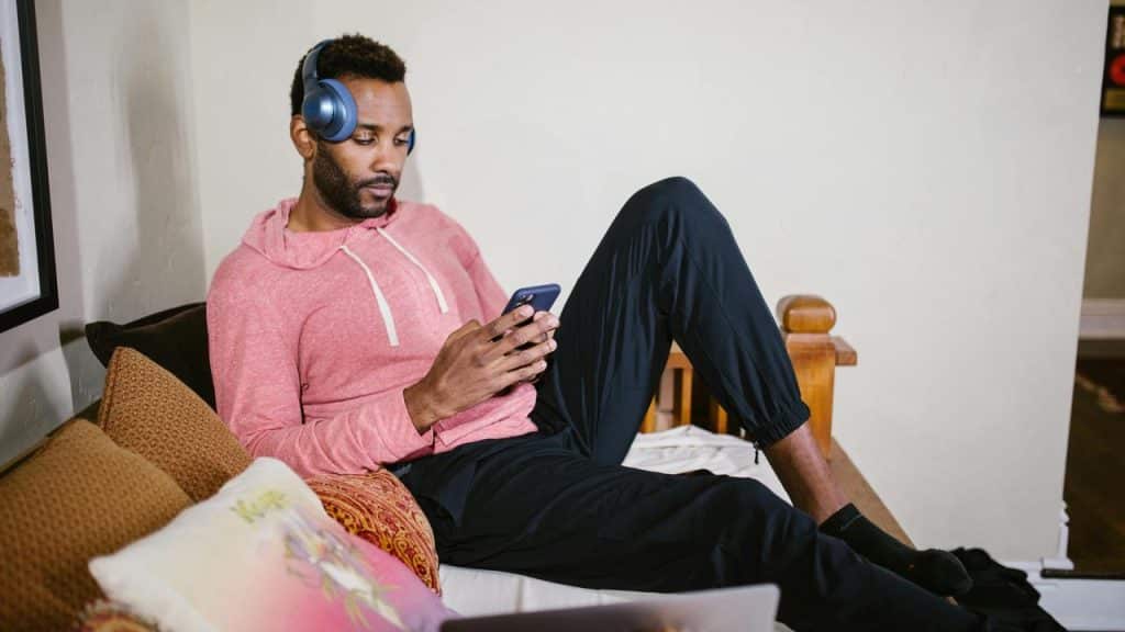 A man wearing headphones and using his phone while relaxing on a bed.