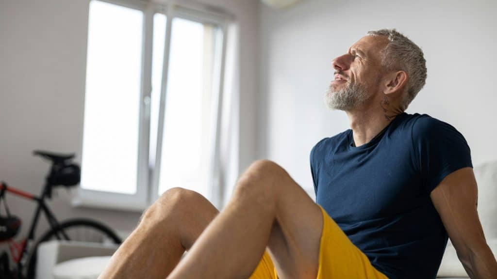 A bearded man in a blue shirt and yellow shorts sits on the floor looking up.