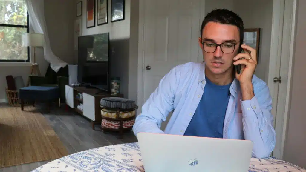A man with glasses talks on a smartphone while sitting at a table with a laptop.