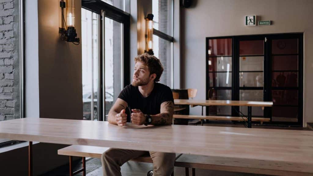 A man sitting alone at a long wooden table in a café looking out the window.