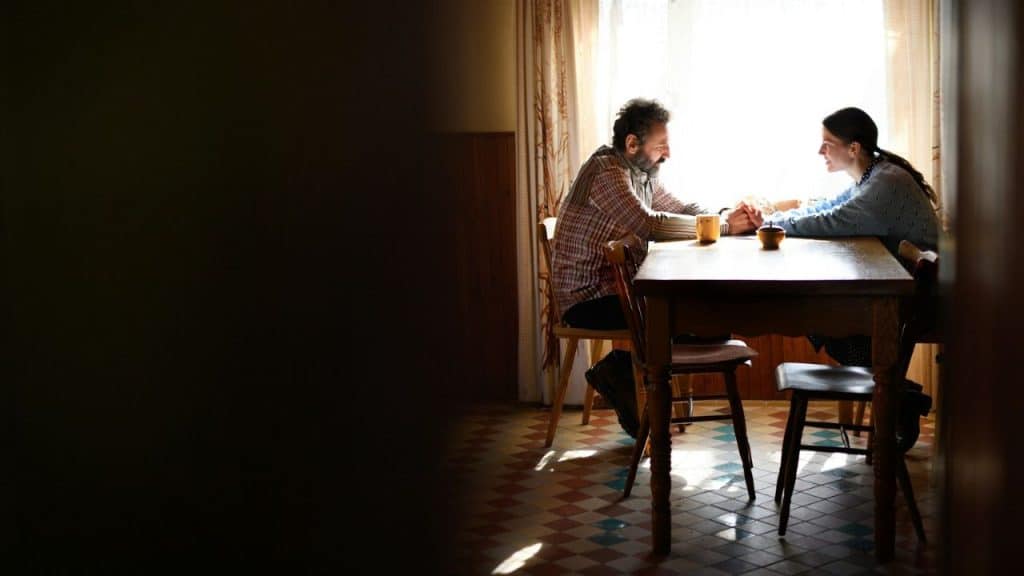 A man and woman sit at a table across from each other, holding hands and looking into each other's eyes, backlit by a window.