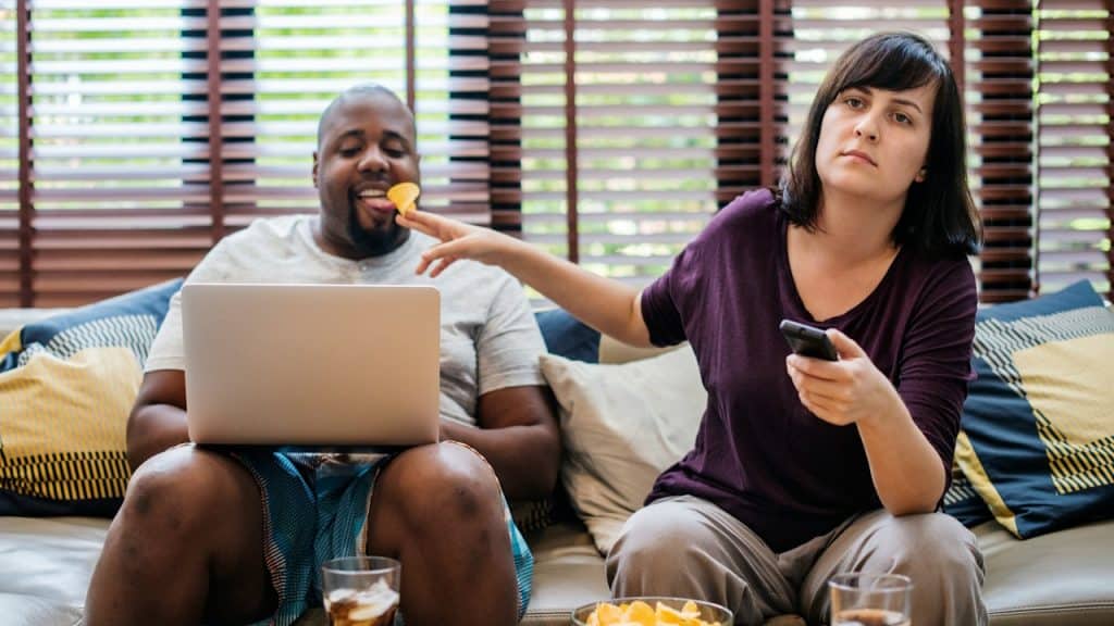 A woman feeding her husband while he is working on his laptop.