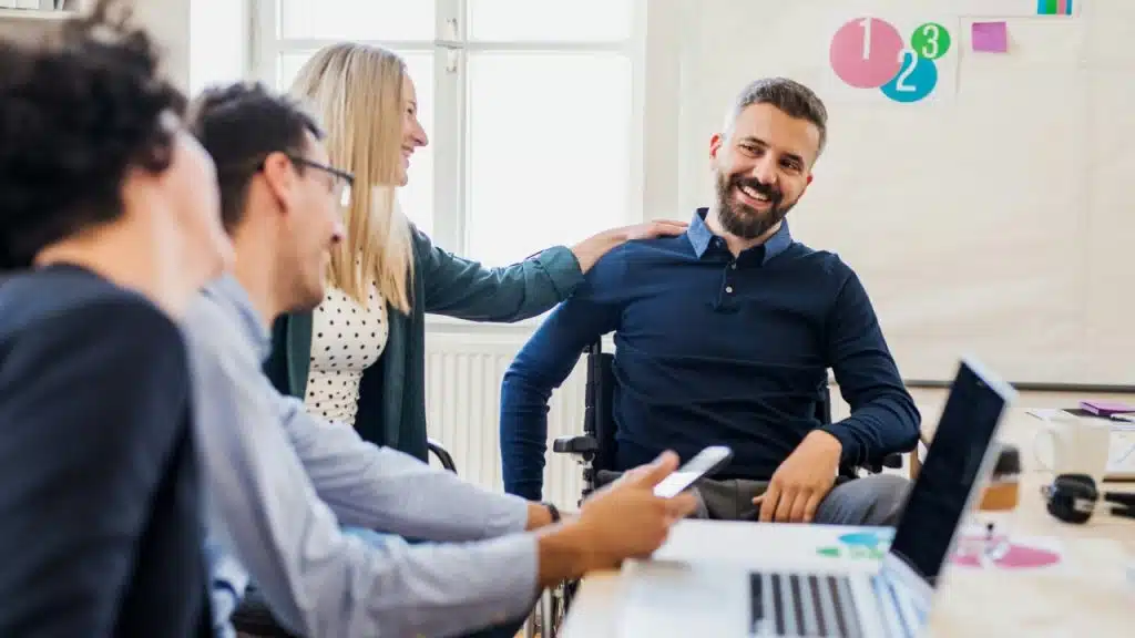 A bearded man in a wheelchair laughs while sitting with colleagues at a bright office table.