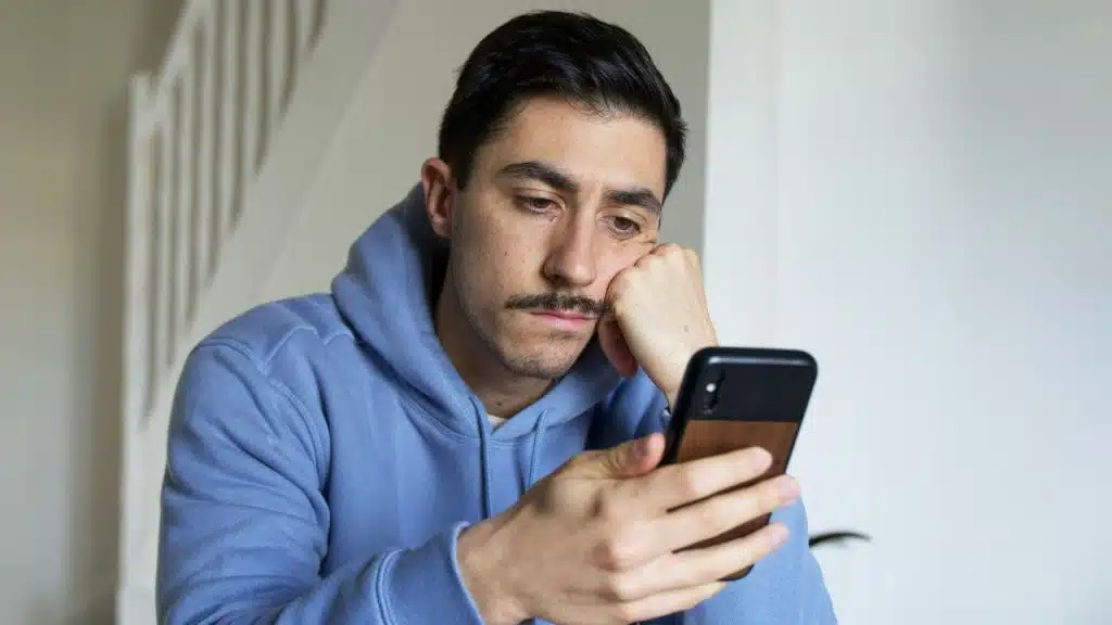 A man with a mustache looks at a mobile phone with a concerned expression.