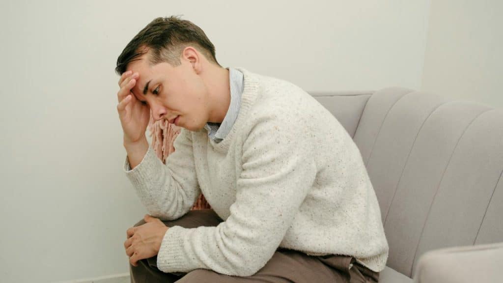 A man sitting on a couch with his hand on his forehead, appearing worried.