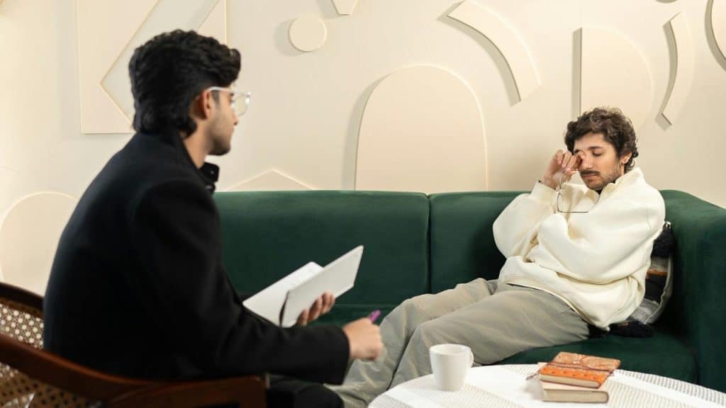 A person rubbing their eyes on a couch opposite someone taking notes.