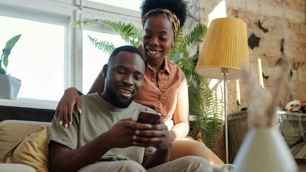 A couple smiling while looking at a phone together in a bright, plant-filled living room.