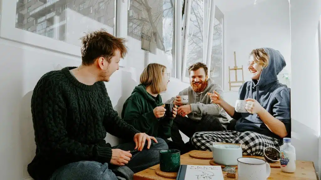 A group of friends laughing and drinking coffee together by a bright window.