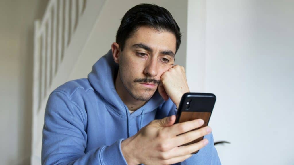 A man with a mustache looks downward with a tired expression while holding a phone.