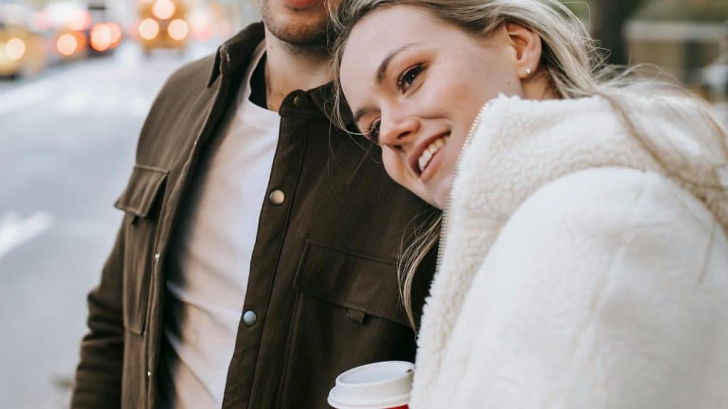 A smiling woman leaning on a man while holding a takeaway coffee on a city street.