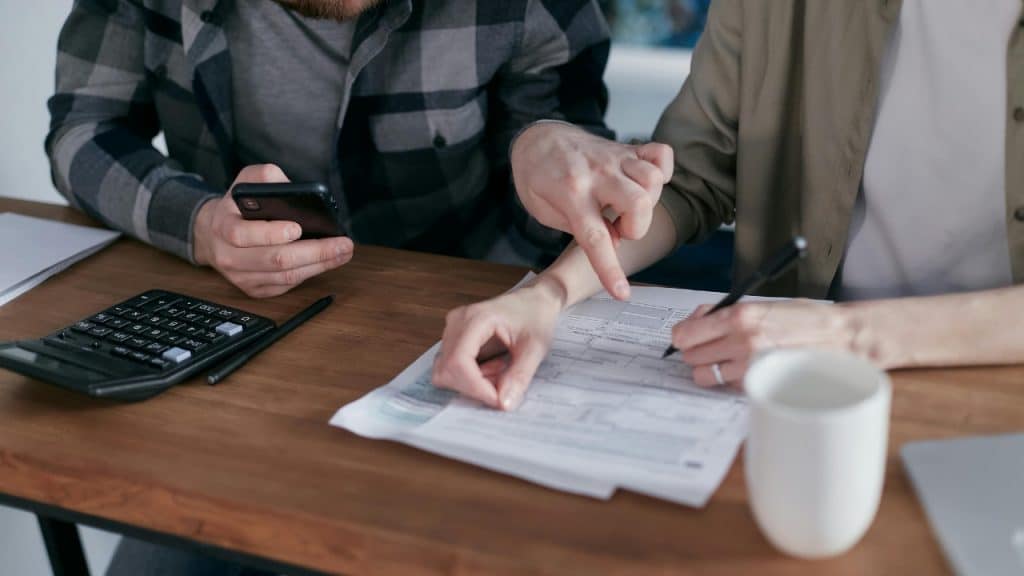 Two people sit at a desk with a calculator, a phone, and a paper document.