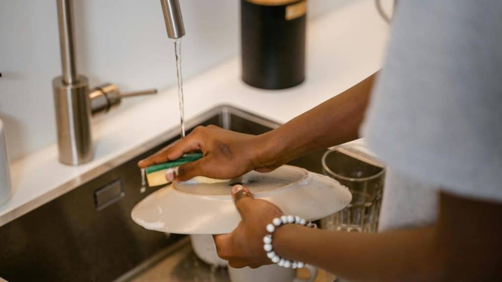 A person washing a plate with a sponge under running water in a kitchen sink.