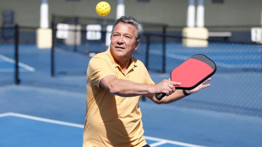 A man in a yellow shirt holds a red paddle while playing on a blue court.