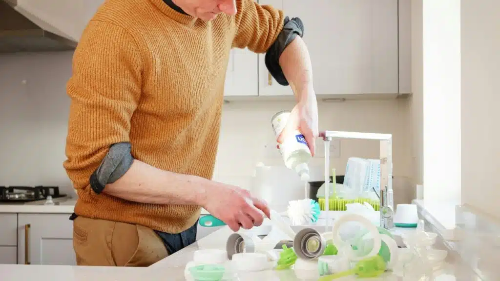 A person in an orange sweater pours liquid soap onto a brush over a sink.