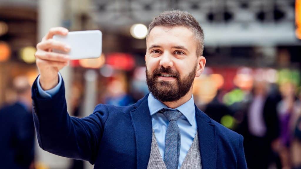 A man in a suit takes a selfie with his phone in a public area.