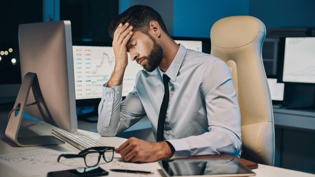 A man in a dress shirt sits at a desk looking tired with his hand on his head.