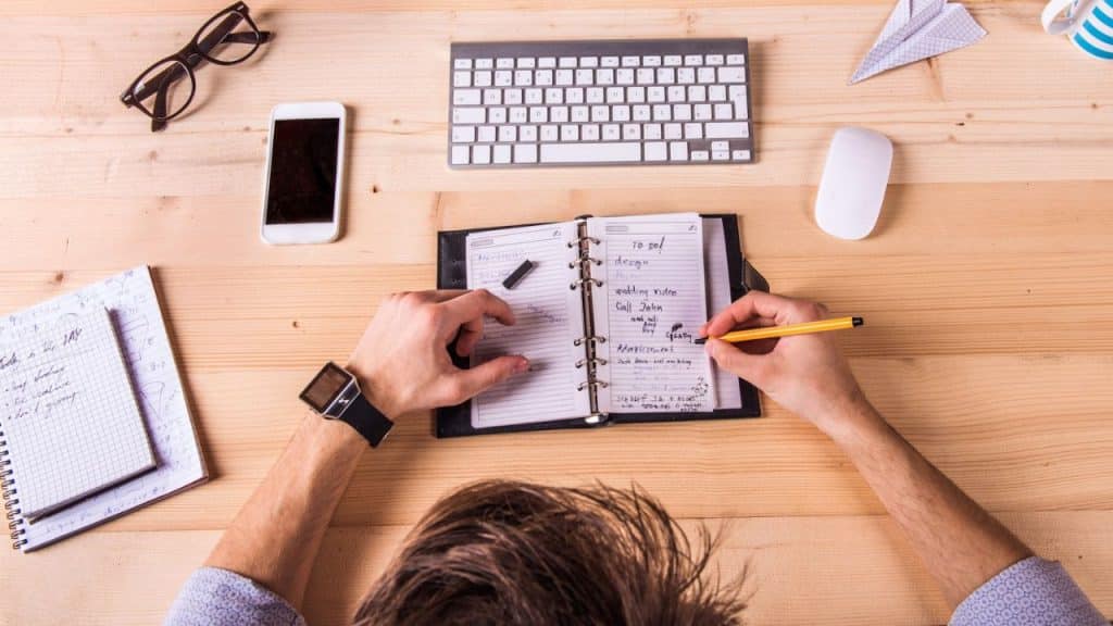A person writes in a notebook at a desk with a keyboard and smartphone.