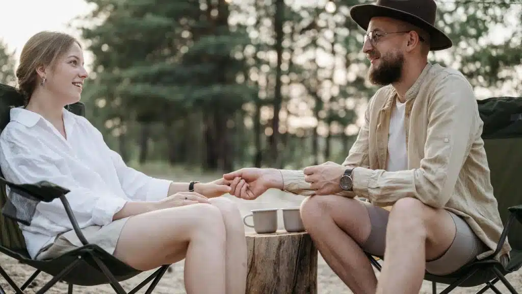 A couple sitting on camping chairs holding hands outdoors.