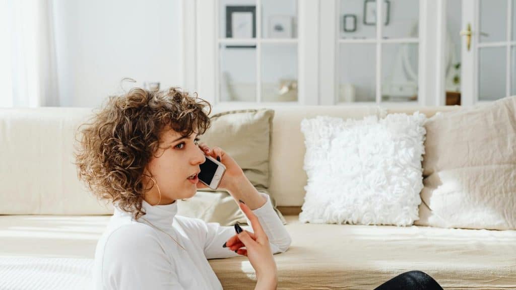 A woman sitting on the floor talking on her smartphone in a bright living room.