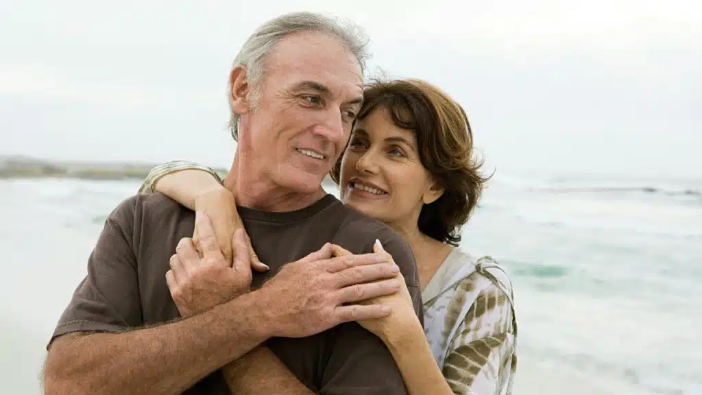 An older couple stands together by the ocean, with the woman embracing the man.