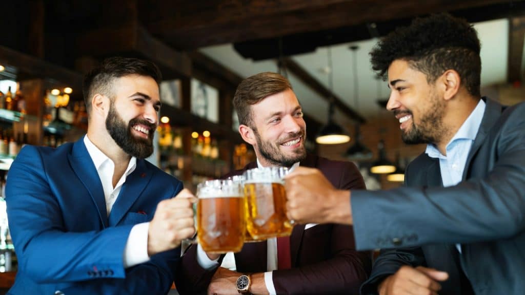 Three men in suits smile and clink glasses of beer together at a wooden bar.