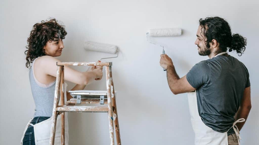 A man and woman use paint rollers to apply white paint to a room wall.